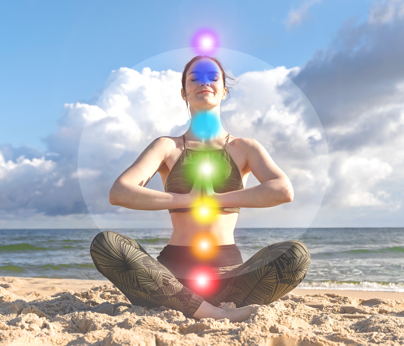 A woman sits meditating on the beach with the 7 chakra colors shown over her body.